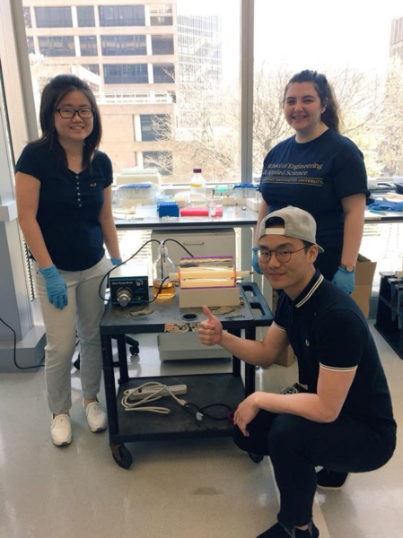 two students standing and one student crouching and giving thumbs up while working in lab